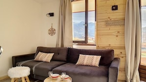 Un salon chaleureux avec un canapé gris foncé, deux coussins à motifs, un petit tabouret blanc et une table ronde en bois sur laquelle reposent deux verres et un bol. On aperçoit un panneau mural en bois avec une fenêtre et une inscription qui dit « PROFITEZ-EN ».