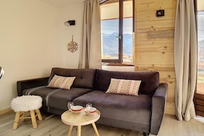 A cozy living room with a dark grey sofa, two patterned cushions, a small white stool, and a round wooden table with two glasses and a bowl. A wooden wall panel with a window and a sign that reads "JUST ENJOY IT" is visible. - 1st floor, view mountain, balcony, tv, ski locker, 26m², Les Menuires (Les Menuires)
