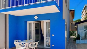 Un edificio azul con balcón, puerta corredera de vidrio y un pequeño patio con mesa y sillas.