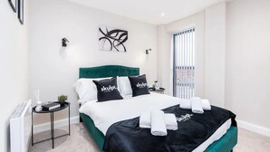 A modern bedroom with a green upholstered headboard, a white and black bedspread, and a view of the city through the window. - Luxury 1 Bed Apt With City Views Heart of Leeds (Leeds)