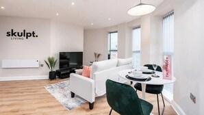 A modern living room with a white sofa, a dining table set for two, a TV mounted on the wall, and a sign reading "skulpt. LIVING". - Luxury 1 Bed Apt With City Views Heart of Leeds (Leeds)