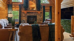 Interior acogedor de una cabaña de troncos con chimenea de piedra, sofás de cuero y un amplio ventanal con vistas a la vegetación.