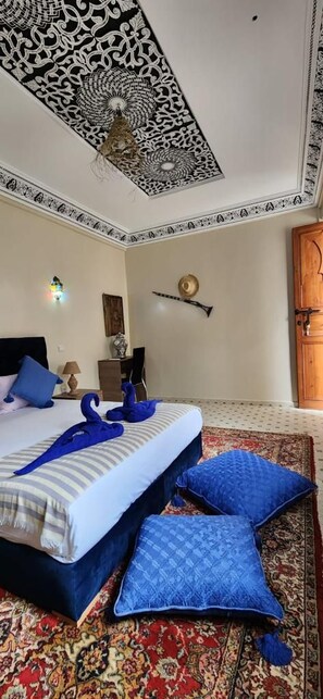 A bedroom with a patterned ceiling, a bed with blue and white bedding, a red patterned rug, and a wooden door.