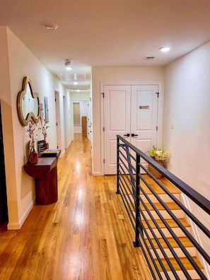 Um corredor com piso de madeira, balcão de recepção, espelho decorativo e escada com corrimão preto.