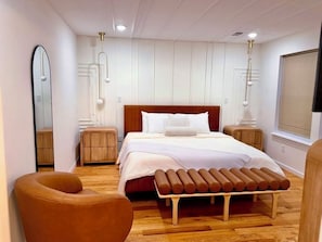 Quarto moderno com piso de madeira, uma cama grande com roupa de cama branca, duas mesas de cabeceira, um espelho e uma janela com persianas.