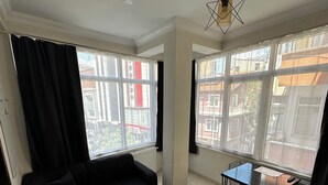 A living room with a black sofa, a dining table with chairs, large windows, and a ceiling fan. - Diva Otel (Isparta)