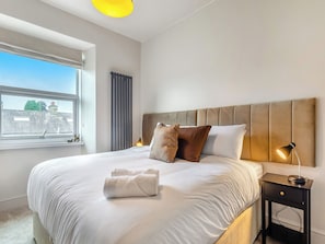 A neatly made bed with pillows, a bedside lamp, and a window with a view of buildings.