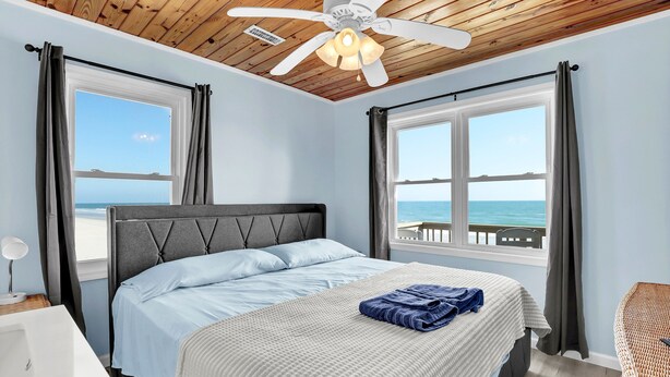 Un dormitorio con cama, ventilador de techo, ventana con vista al mar y mesita de noche con lámpara.