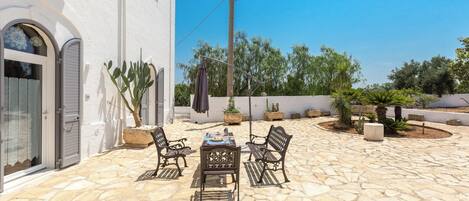 A stone patio with a table and chairs, surrounded by potted plants and a white building with arched windows.