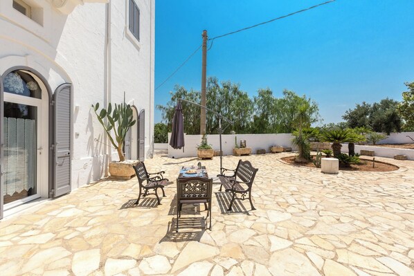 A stone patio with a table and chairs, surrounded by potted plants and a white building with arched windows.