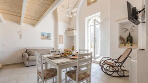 A dining area with a white table set for four, surrounded by white chairs with floral cushions. A wooden rocking chair is positioned near a white wall with a mounted television.