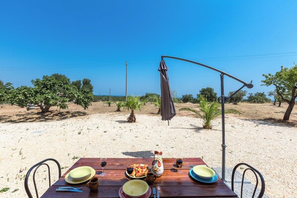 A wooden table set for a meal with plates, glasses, and a ceramic jug, overlooking a desert landscape with sparse vegetation and a clear sky.