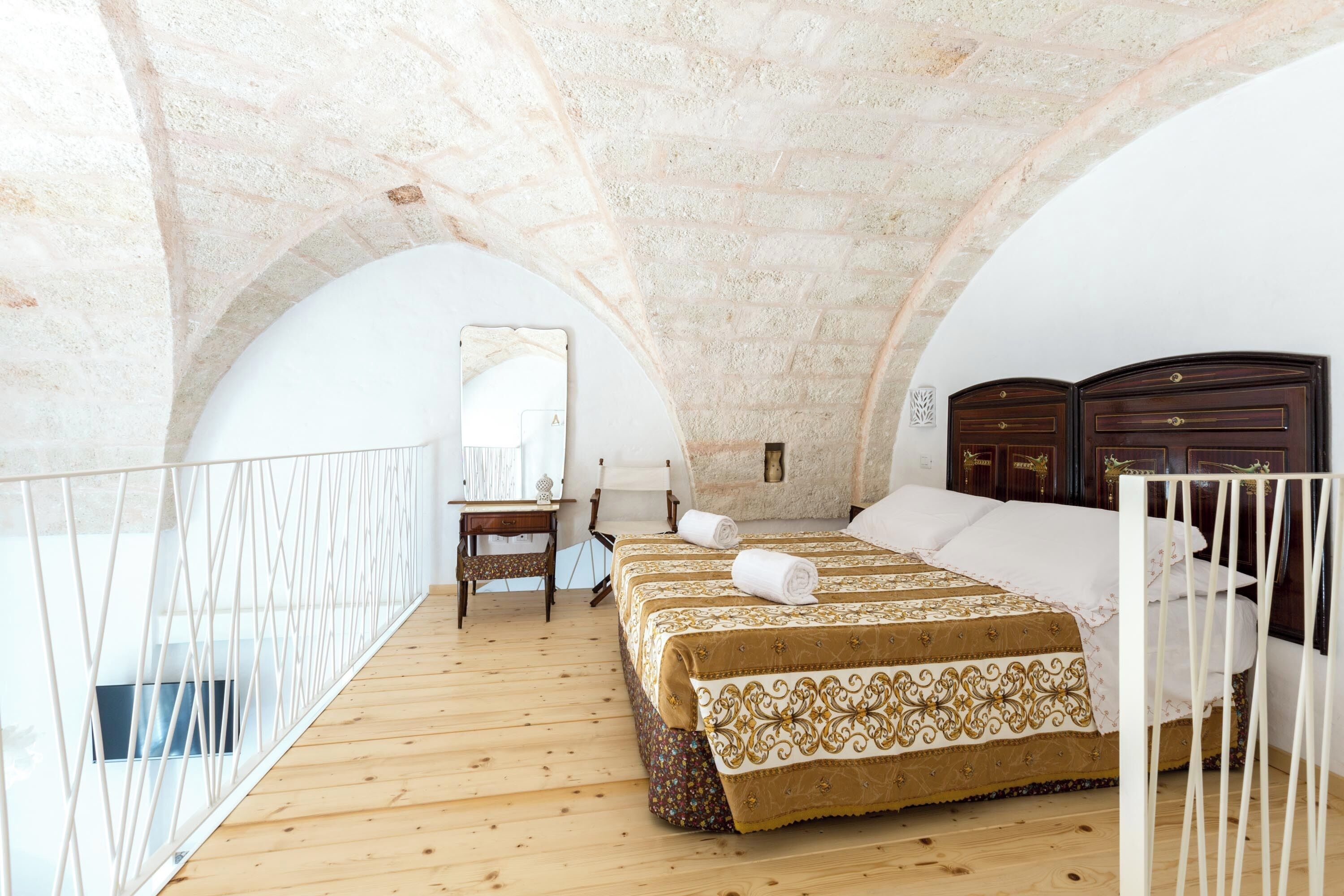 Quarto com piso de madeira, cama com colcha estampada, mesa de cabeceira, espelho e guarda-roupa de madeira.