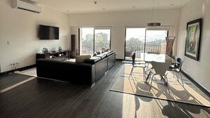 Un vaste espace de vie doté d’un parquet en bois foncé, d’un canapé noir, d’une table à manger avec des chaises et d’une grande fenêtre offrant une vue sur la ville.