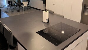 Une cuisine moderne dotée d’une plaque de cuisson à induction, de luminaires suspendus et d’un coin repas avec une table et des chaises en verre.