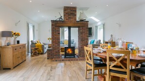 A cozy living room with a brick fireplace, wooden furniture, and a dining area with a table set for breakfast. - Bwthyn Alberts -  an on the beach that sleeps 7 guests  in 3 bedrooms (Trearddur Bay)
