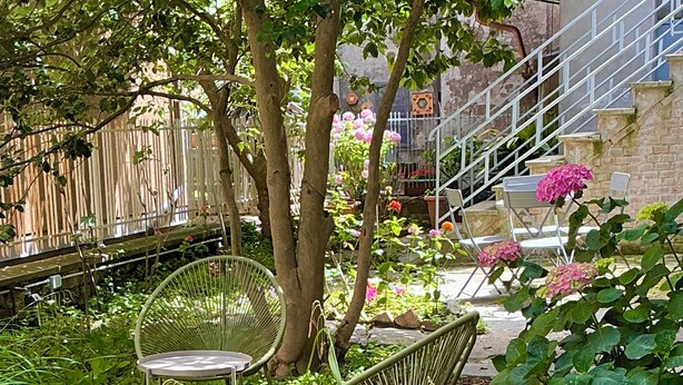 A garden with a metal chair, a wicker chair, and a bench. There are trees and plants, including a pink flowering plant. - Suite Cavallotti - The Almond (Orvieto)