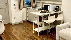 A compact kitchen with a microwave, toaster, and kettle, a small dining table with chairs, a window with curtains, and a white door. - Suite Cavallotti - The Almond (Orvieto)