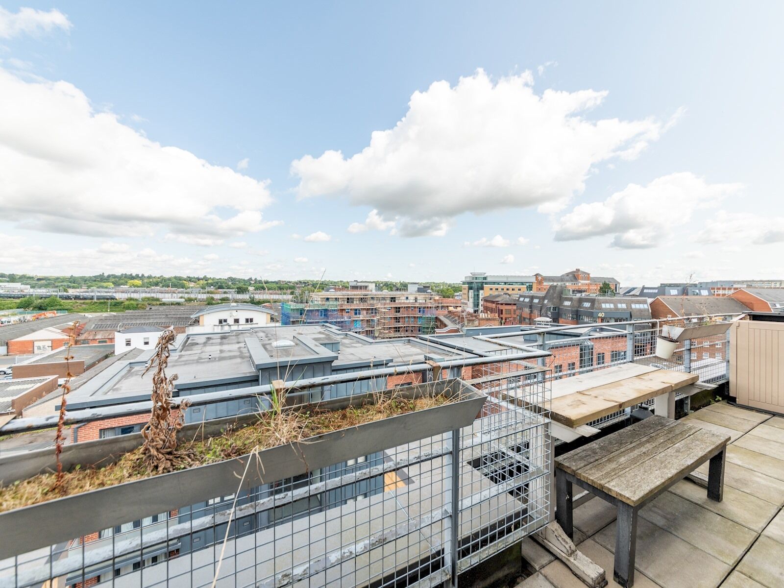 A rooftop terrace with a view of surrounding buildings, a metal fence, and a wooden bench.