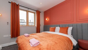 Une chambre d’hôtel avec un grand lit, une literie orange, une tête de lit grise et une vue sur les bâtiments par la fenêtre.