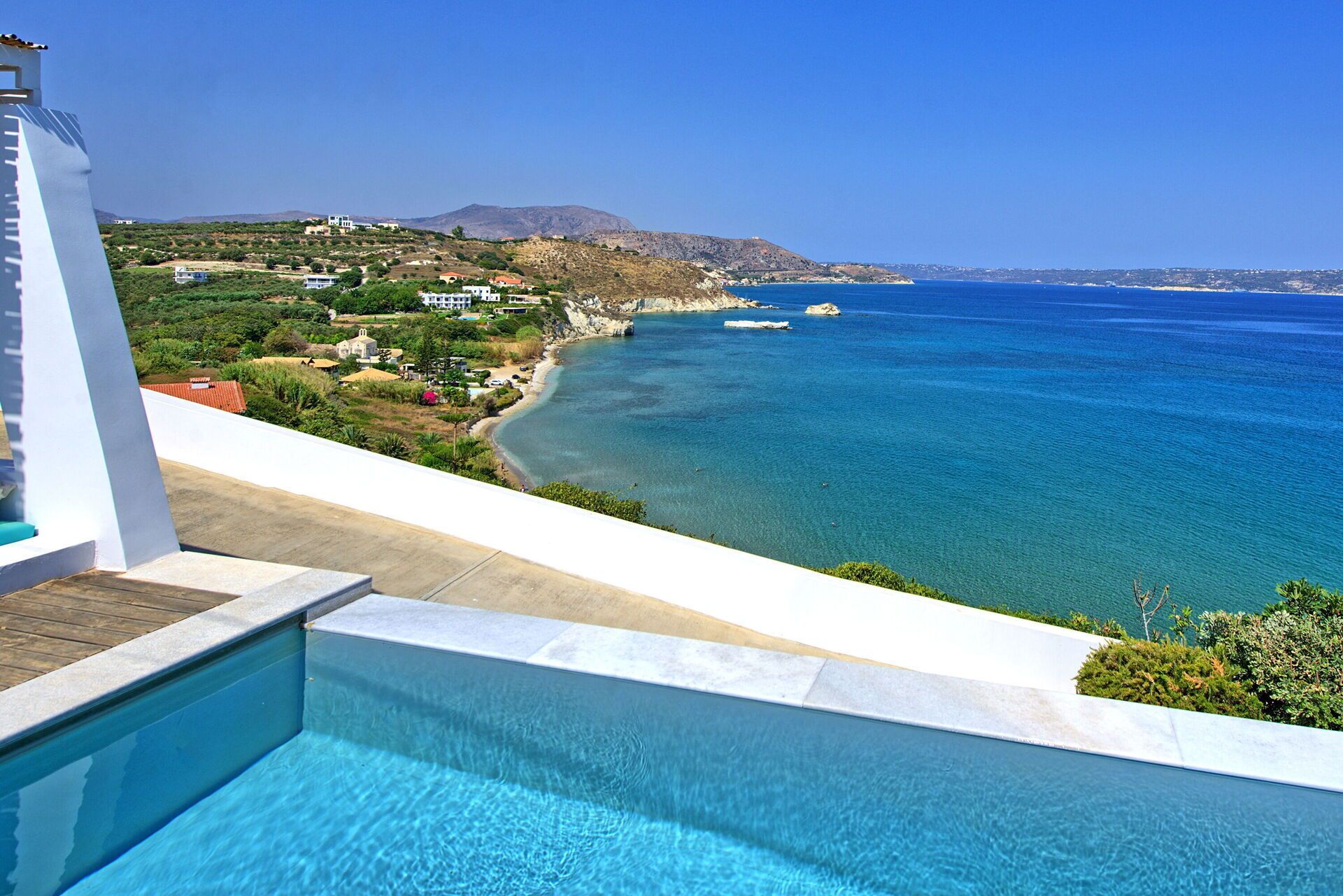 A poolside view overlooking a coastal landscape with clear blue waters, a beach, and distant hills.