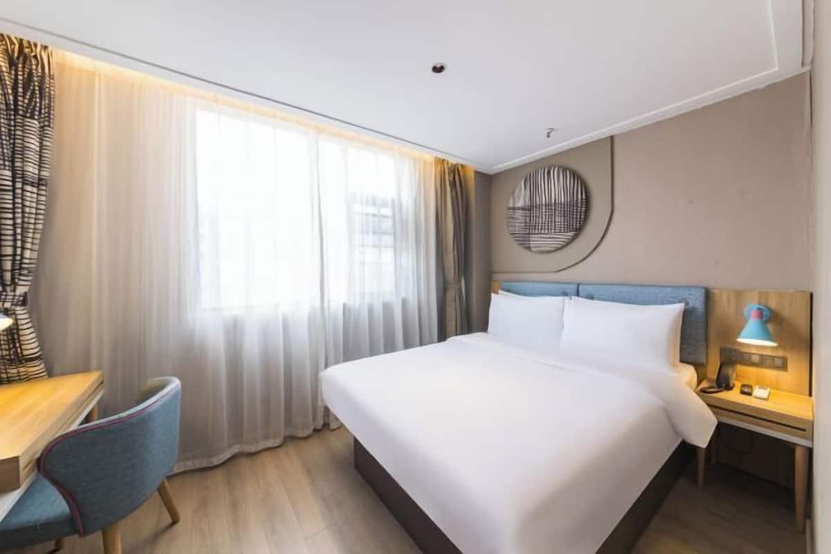 A hotel room with a bed, a desk, a chair, a nightstand, and a lamp.