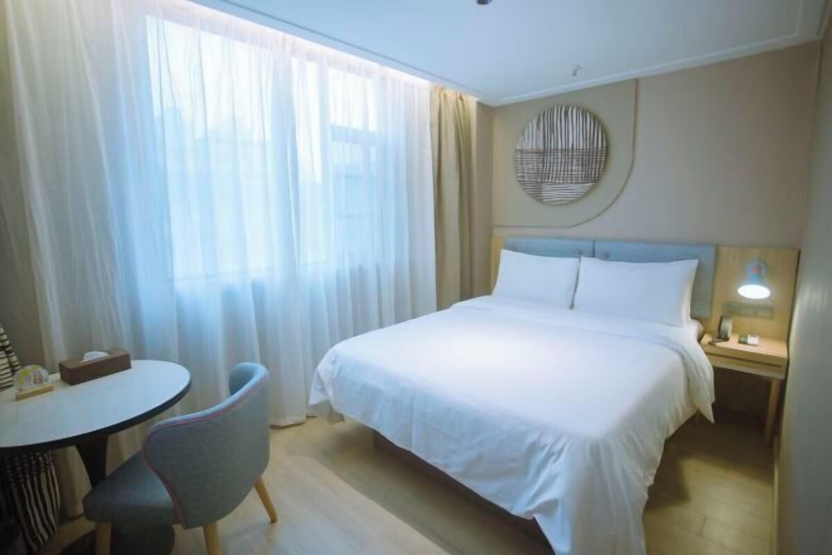 A hotel room with a bed, a bedside table with a lamp, a chair, a round table, and a window with curtains.