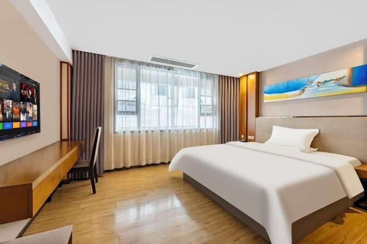 A modern hotel room with a large bed, a flat-screen TV mounted on the wall, a wooden desk, and a large window with curtains.