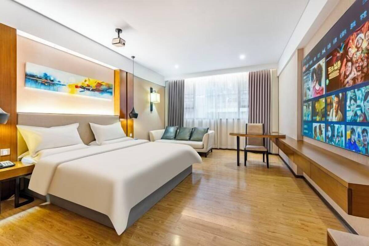 A modern hotel room with a large bed, a desk, a chair, a TV wall, and a window with curtains.