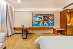 Quarto de hotel com cama, escrivaninha, cadeira, parede com TV e várias imagens, e um armário de madeira.