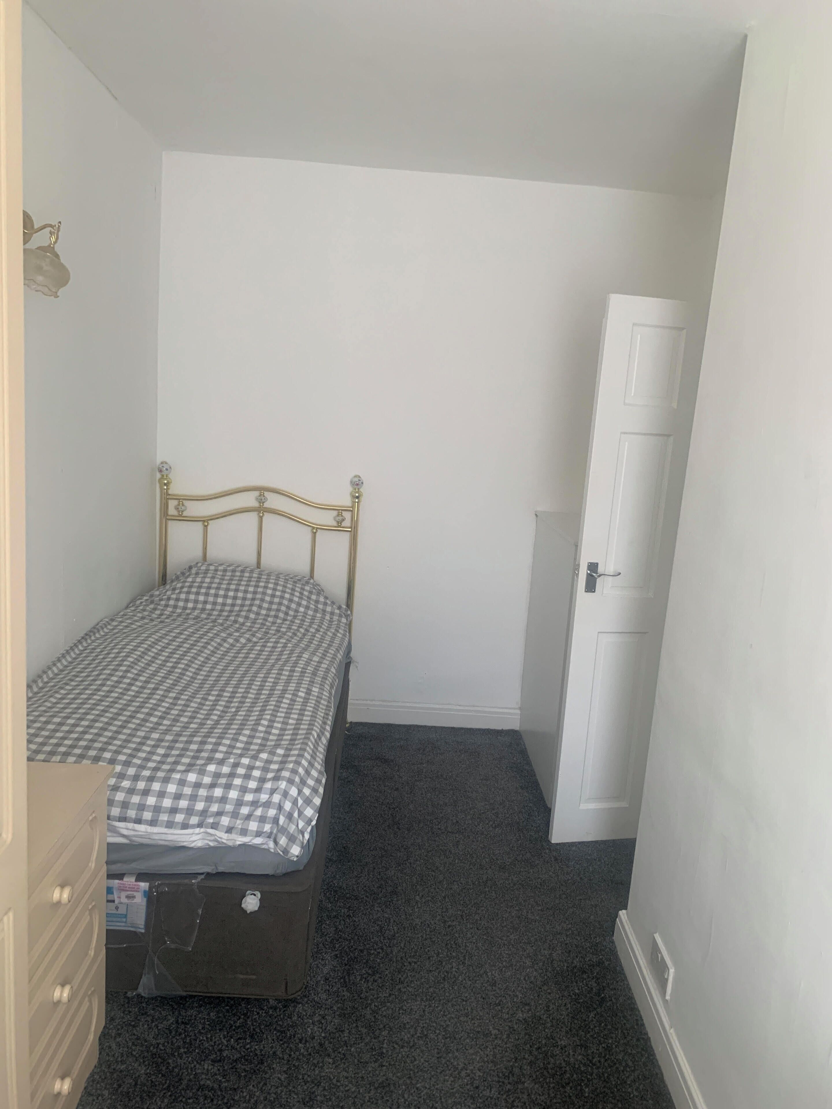 A small, sparsely furnished room with a single bed, a dresser, and a door.