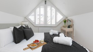 Ein Schlafzimmer mit geneigter Decke, einem Bett mit schwarz-weiß gestreifter Bettwäsche, einem Nachttisch mit Lampe, einer kleinen Holzkommode und einem Fenster mit weißen Fensterläden.