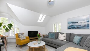 Ein modernes Wohnzimmer mit Sofa, Sessel und Couchtisch. Es gibt einen Fernseher, einen Topfpflanzen und ein Fenster mit Jalousien.