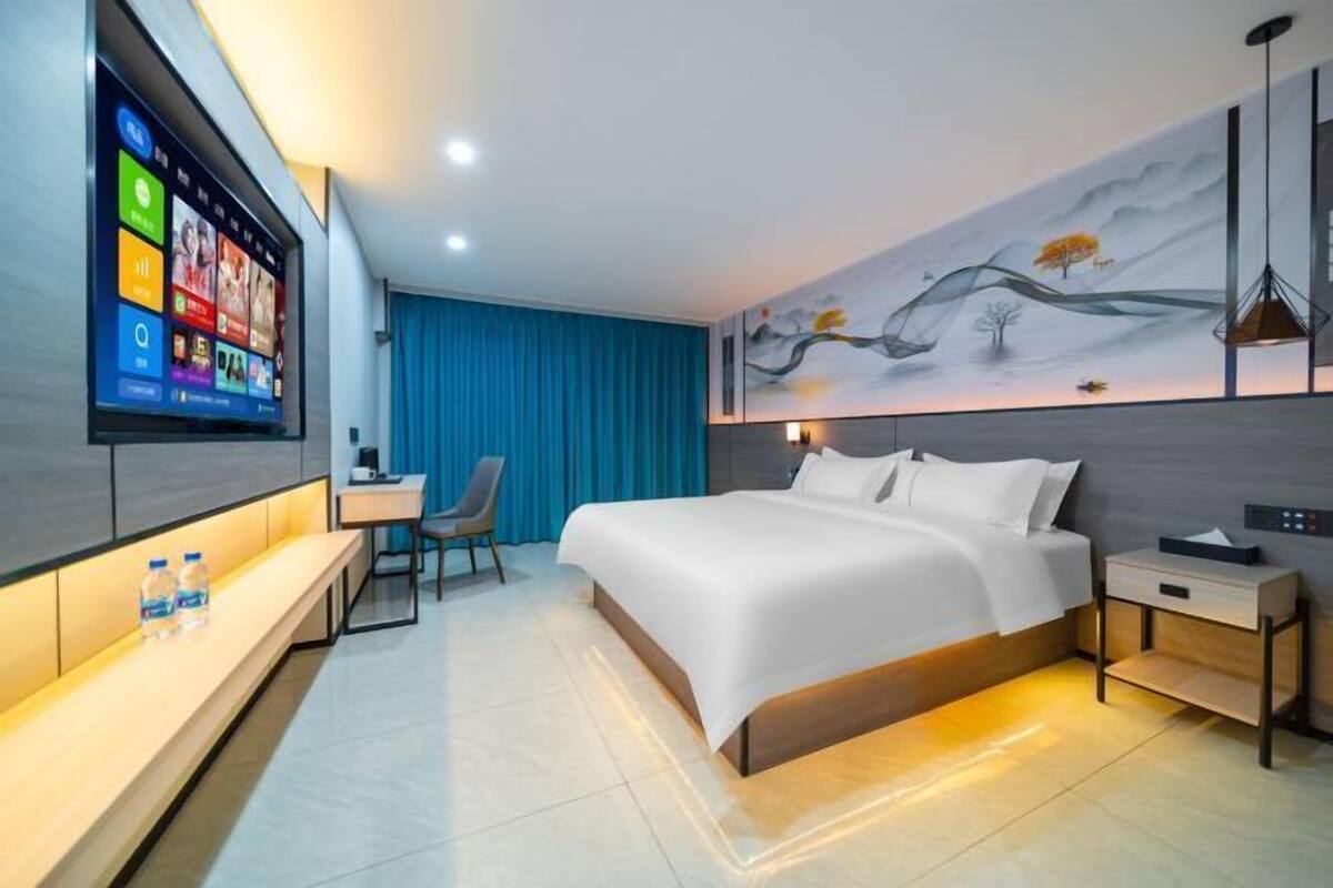 A modern hotel room with a large bed, a desk, a chair, a TV mounted on the wall, and a painting on the wall.
