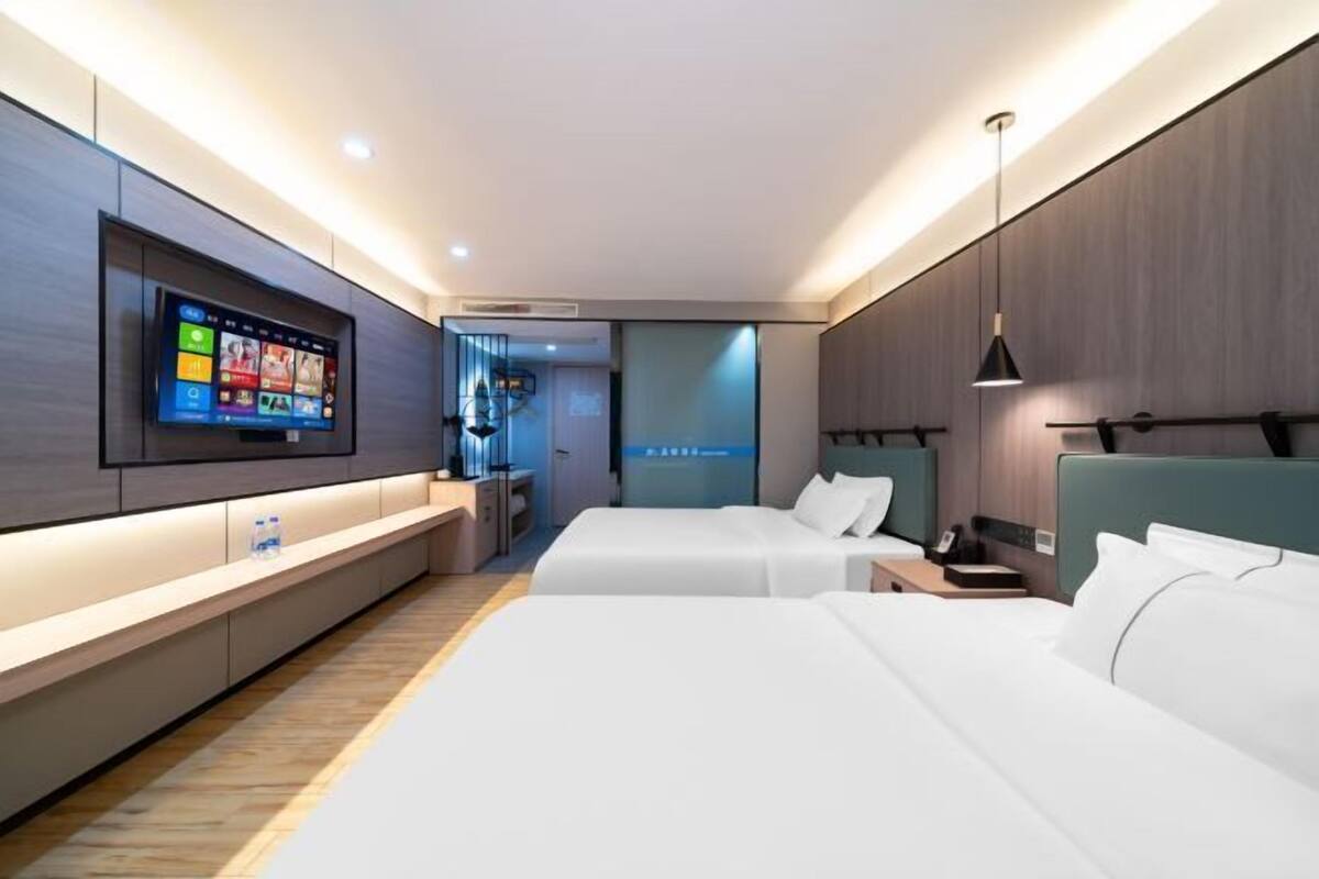A modern hotel room with two beds, a flat-screen TV mounted on the wall, a bedside table, and a small seating area.
