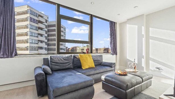 A modern living room with a grey sectional sofa, a matching ottoman, and a large window offering a view of a cityscape. - Heathrow Retail Park Apartment Sleeps SEVEN Turbo Wi-Fi (Shepperton)