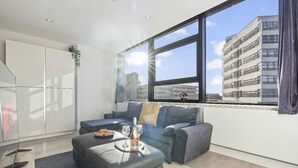 A modern living room with a large window, a sofa, a coffee table with a bottle and glasses, and a view of a building. - Heathrow Retail Park Apartment Sleeps SEVEN Turbo Wi-Fi (Shepperton)