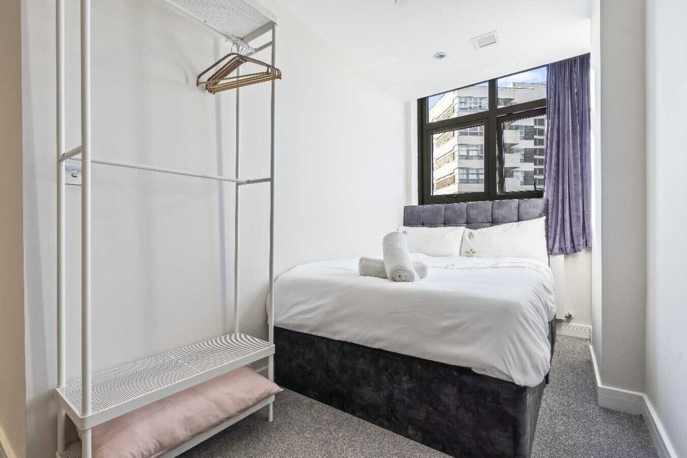Une chambre avec un lit, une armoire blanche, une douche suspendue et une fenêtre donnant sur des immeubles.