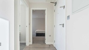 A narrow hallway with a wooden floor leading to a bedroom with a bed and a dark-colored chest of drawers. - Heathrow Retail Park Apartment Sleeps SEVEN Turbo Wi-Fi (Shepperton)