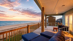 Una terraza de madera con vistas a la playa y al océano, un espacio de descanso cubierto con cojines, y un edificio con amplios ventanales.