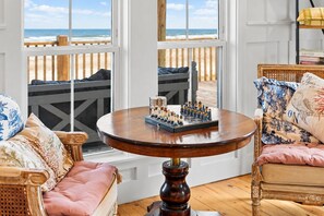 A room with a wooden deck overlooking the ocean, a round wooden table with chess pieces, and two wicker chairs with cushions. - Beachfront Home | Direct Beach Access | Gulf Views | Large Deck | Pet Friendly (Gilchrist)
