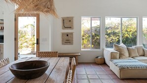 Una habitació amb una taula de dinar de fusta, una làmpada de sostre teixida, un sofà amb coixins estampats i vista a arbres a través de grans finestres.