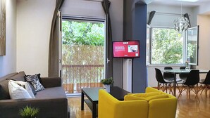 Un saló modern amb un sofà, cadires grogues, una taula de menjador amb cadires, un televisor i una finestra gran amb vista a vegetació.