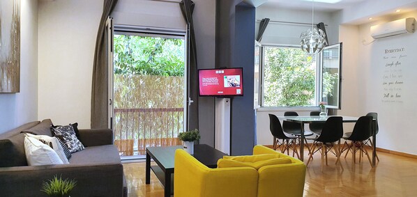 En moderne stue med sofa, gule stole, spisebord med stole, fjernsyn og et stort vindue med udsigt til grøn natur.
