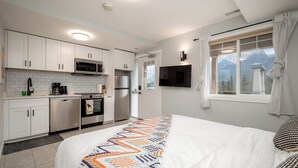 Una cocina moderna con armarios blancos, un microondas, una estufa y un refrigerador. Hay un televisor montado en la pared y una ventana con cortinas.