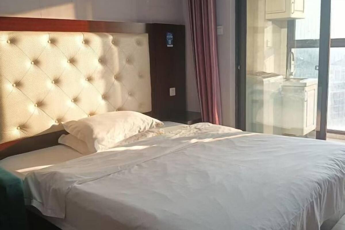 A hotel room with a tufted headboard, a large bed with white bedding, and a view of the city through a glass door.