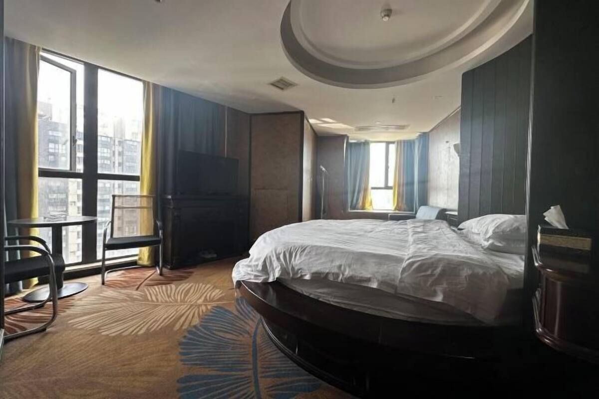 A hotel room with a large bed, a TV, a desk, and a view of the city through large windows.