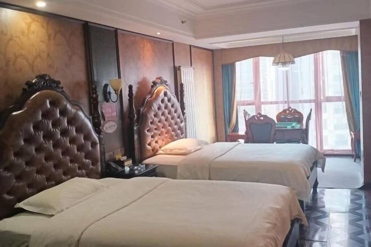 A hotel room with two beds, a wooden headboard, a nightstand, a desk, and a view of the city through large windows.