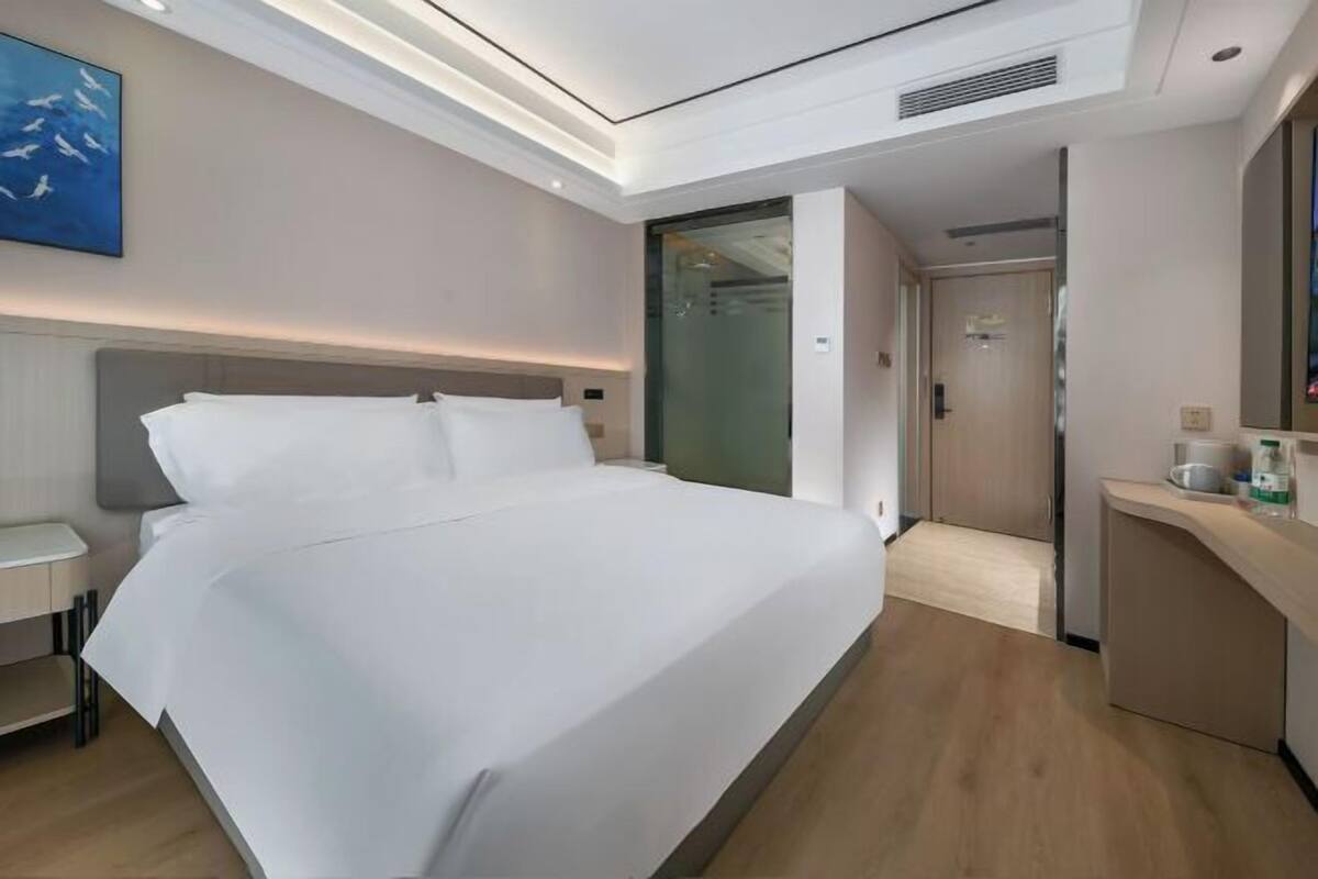 Ein modernes Hotelzimmer mit einem großen Bett, einem Nachttisch, einem Schreibtisch und einem durch eine Glastür sichtbaren Badezimmer.