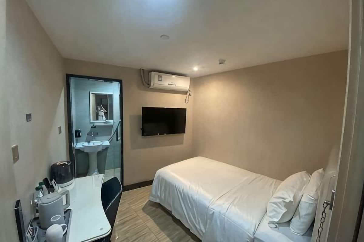 Ein Hotelzimmer mit Bett, Schreibtisch mit Stuhl, Waschbecken und einem an der Wand befestigten Fernseher.